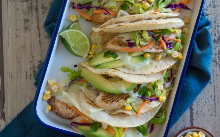 Three fish tacos with avocado, lettuce, cabbage, carrot, and corn, garnished with lime wedges and creamy sauce, served in a tray on a wooden table.