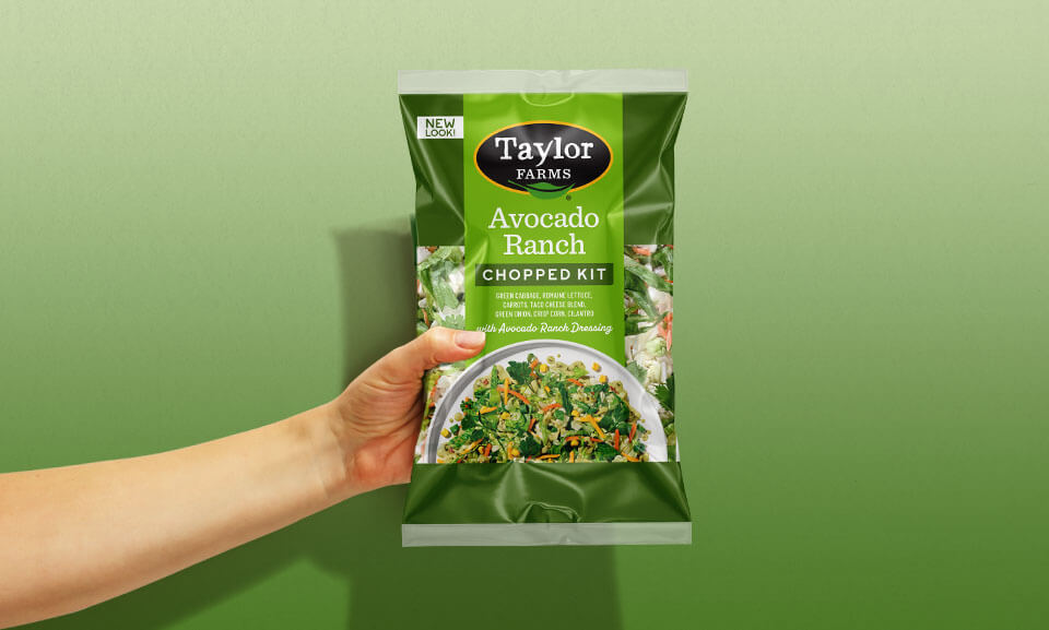 Hand holding a Taylor Farms Avocado Ranch Chopped Salad Kit bag featuring fresh ingredients and vibrant packaging.