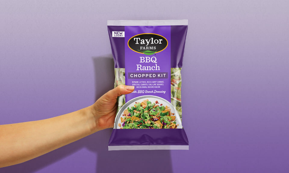 Hand holding a bag of Taylor Farms BBQ Ranch Chopped Salad Kit, showcasing fresh ingredients and vibrant packaging.