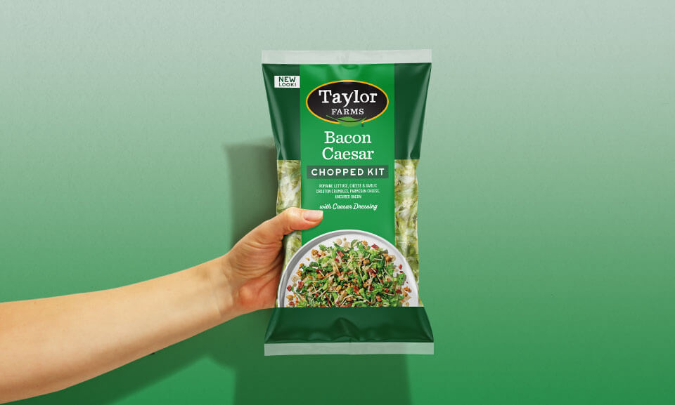 Hand holding a bag of Taylor Farms Bacon Caesar Chopped Salad Kit, showcasing fresh ingredients and packaging design.