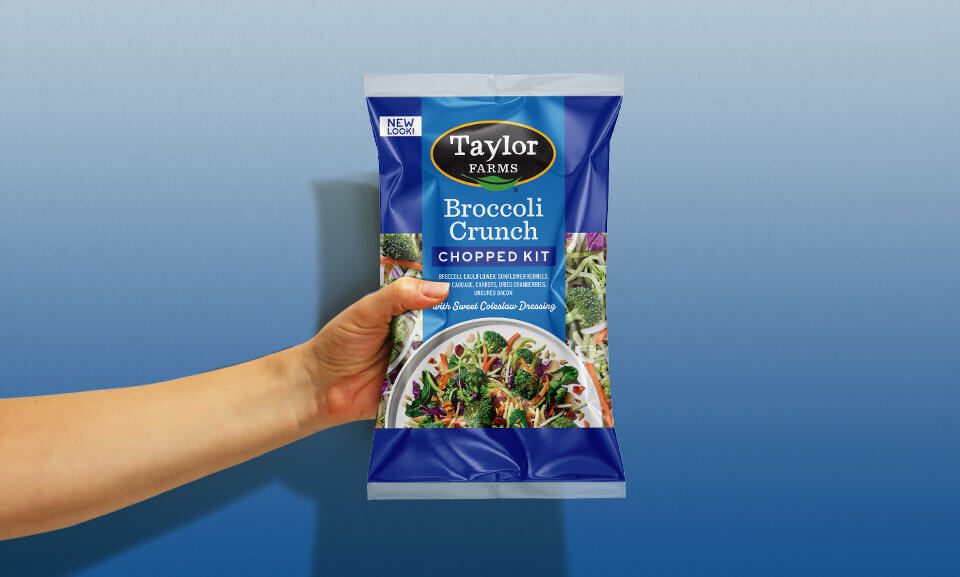 Hand holding a bag of Taylor Farms Broccoli Crunch Chopped Salad Kit, showcasing fresh ingredients and vibrant packaging.