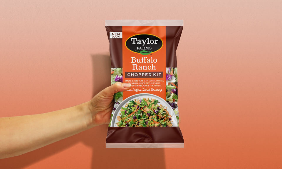 Hand holding a bag of Taylor Farms Buffalo Ranch Chopped Salad Kit, showcasing fresh ingredients and convenient packaging.