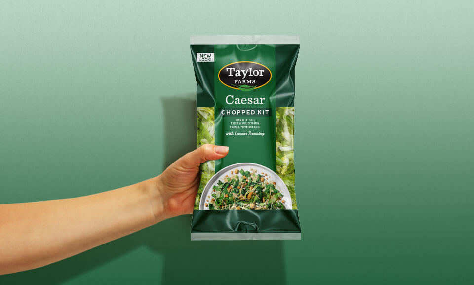 Hand holding a bag of Taylor Farms Caesar Chopped Kit featuring fresh salad ingredients and flavorful toppings.