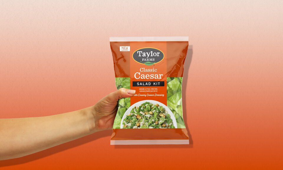 A person holding a bag of Taylor Farms Classic Caesar Salad Kit ready for fresh meal preparation.