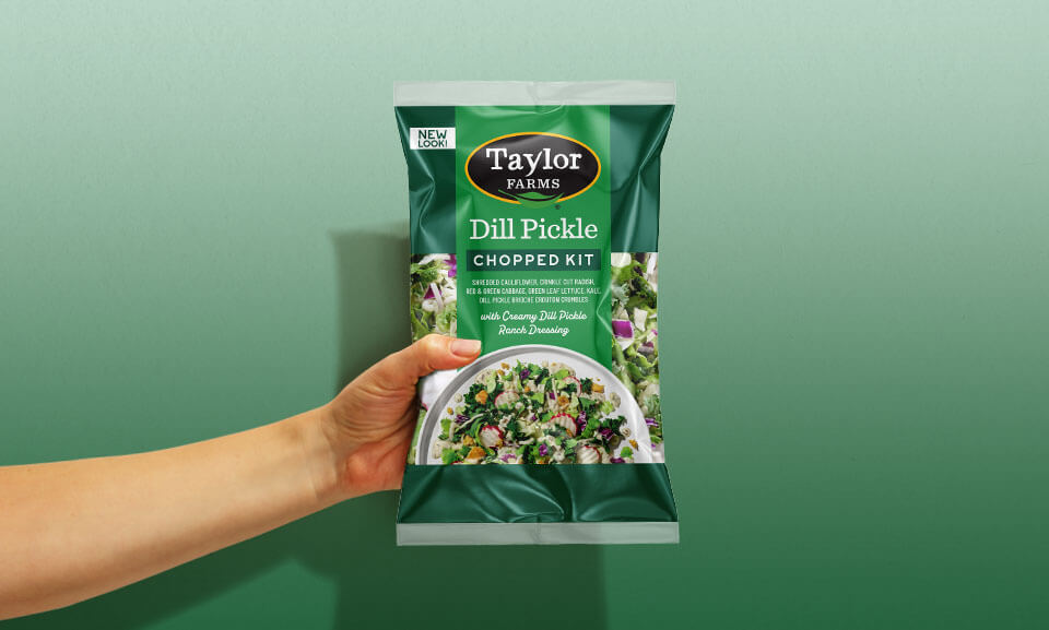 Hand holding a bag of Taylor Farms Dill Pickle Chopped Salad Kit, showcasing its vibrant ingredients and packaging.