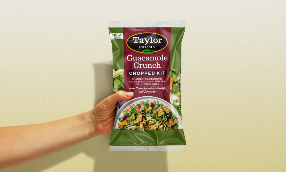Hand holding a bag of Taylor Farms Guacamole Crunch Chopped Kit, showcasing fresh ingredients for a healthy salad.