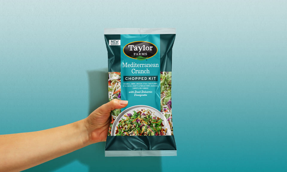Hand holding a bag of Mediterranean Crunch Chopped Kit from Taylor Farms, showcasing fresh ingredients for a healthy meal.