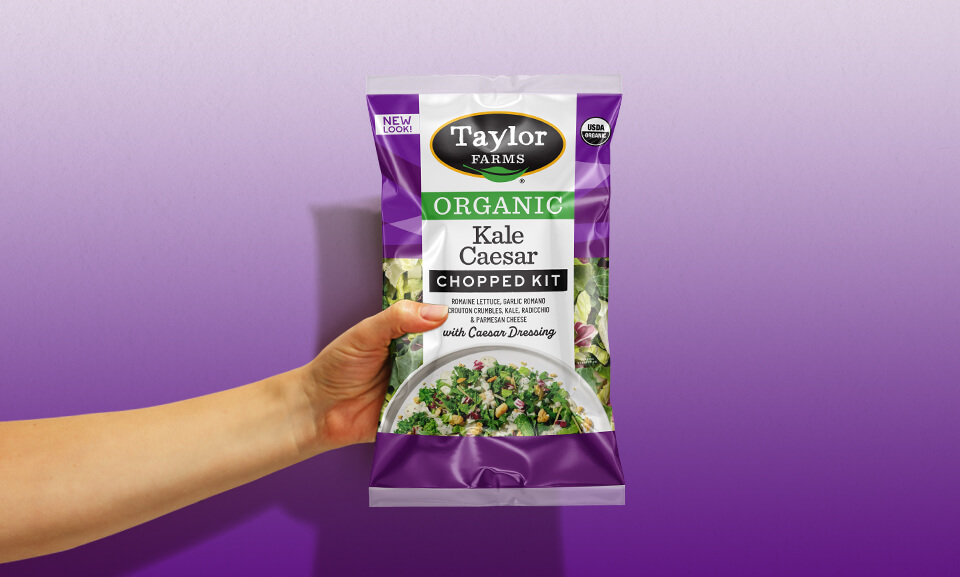 Person holding a bag of Taylor Farms Organic Kale Caesar Chopped Kit, showcasing fresh salad ingredients.