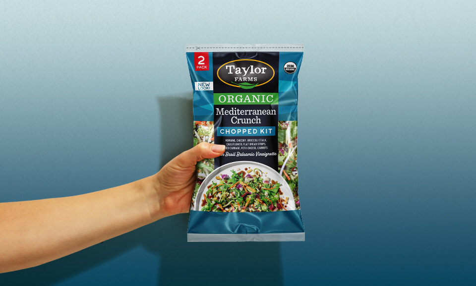 Hand holding a bag of Taylor Farms Organic Mediterranean Crunch Chopped Salad Kit, showcasing vibrant ingredients.