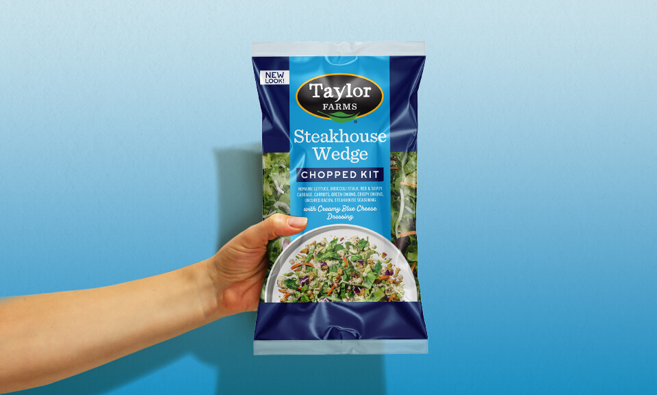 Hand holding a bag of Taylor Farms Steakhouse Wedge Chopped Kit with fresh ingredients visible.