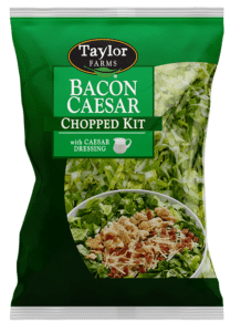 The Bacon Caesar Chopped Salad kit package, showing chopped romaine lettuce topped with cheese garlic crouton crumbles, Parmesan cheese, bacon, and tangy Caesar dressing.