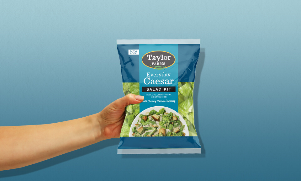 Fresh Taylor Farms Everyday Caesar Salad Kit being held with vibrant greens and croutons visible in the packaging.