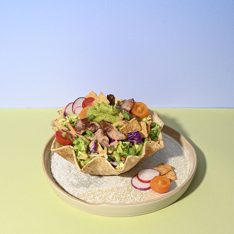 The Guacamole Crunch Chopped Salad Kit in a taco bowl, showing green leaf lettuce, shredded broccoli, red & savoy cabbage, carrots, and green onion, which are topped off with real guacamole, salsa picante dressing, and chili lime tortilla chips.