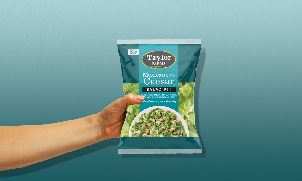 Hand holding a bag of Taylor Farms Mexican Style Caesar Salad Kit, showcasing fresh ingredients and packaging design.