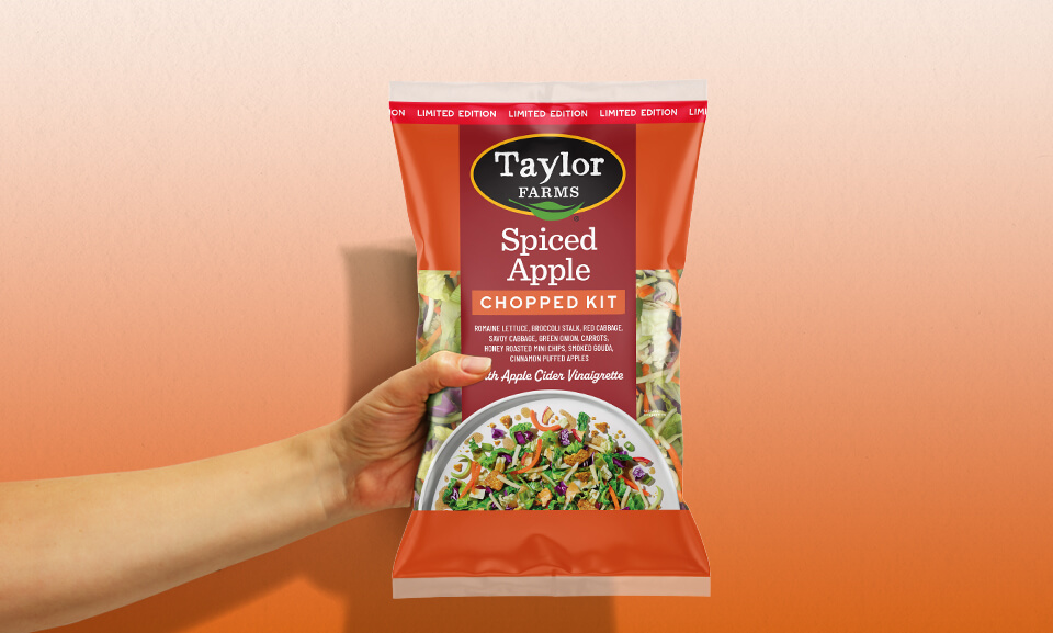 Hand holding a bag of Taylor Farms Spiced Apple Chopped Kit, showcasing fresh ingredients and vibrant packaging.