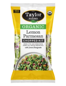 Taylor Farms Organic Lemon Parmesan Chopped Salad package, next to an open tortilla on a plate with the Lemon Parmesan Salad, topped with sliced avocado and a creamy sauce.