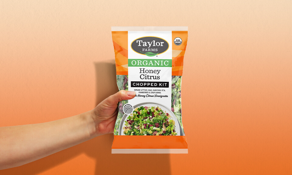 Hand holding a bag of Taylor Farms Organic Honey Citrus Chopped Salad Kit with vibrant ingredients visible.