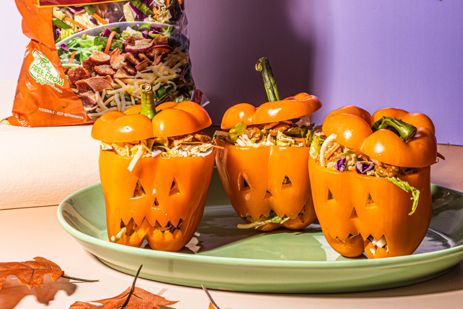 Three stuffed orange bell peppers on a plate, which have been carved into jack-o-lanterns, with a Spiced Apple Chopped Salad Kit in the background.