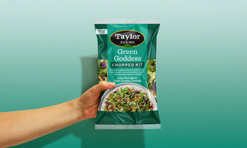 Hand holding a bag of Taylor Farms Green Goddess Chopped Salad Kit, showcasing fresh ingredients and vibrant packaging.