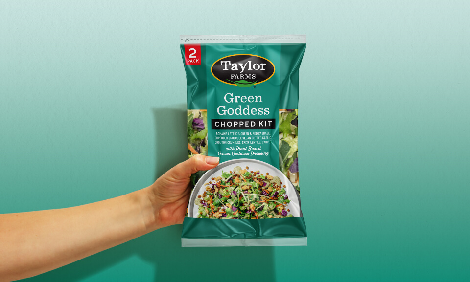 Hand holding a bag of Taylor Farms Green Goddess Chopped Salad Kit, showcasing fresh ingredients and vibrant packaging.