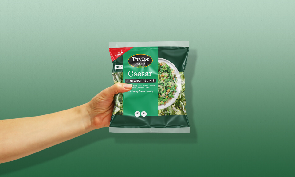 Hand holding a bag of Taylor Farms Caesar Mini Chopped Salad Kit, showcasing fresh ingredients and vibrant packaging.