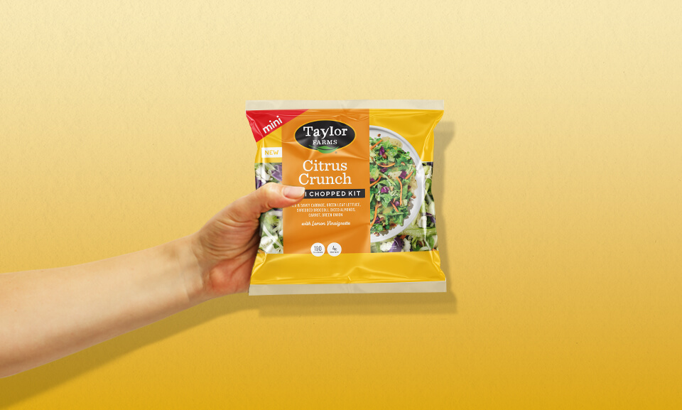 Hand holding a bag of Taylor Farms Citrus Crunch Mini Chopped Salad Kit with vibrant packaging.