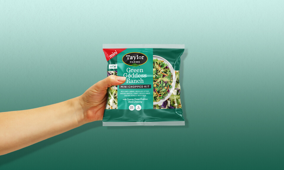 Hand holding a bag of Taylor Farms Green Goddess Mini Chopped Salad Kit, fresh ingredients visible.