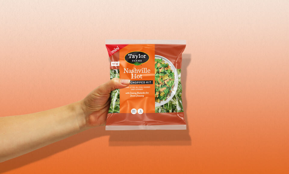 Hand holding a Nashville Hot Mini Chopped Kit bag from Taylor Farms, featuring fresh ingredients and vibrant packaging.
