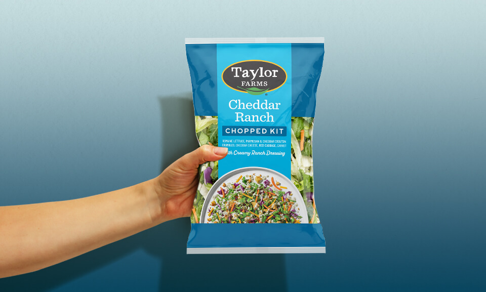Hand holding a Cheddar Ranch Chopped Salad Kit bag from Taylor Farms, showcasing fresh ingredients and packaging design.