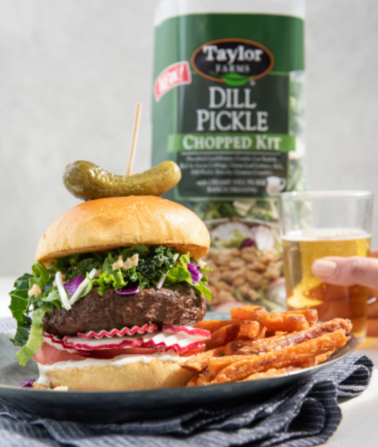Dill pickle burger topped with fresh ingredients, nestled on a wooden board, showcasing Taylor Farms' gourmet offerings.