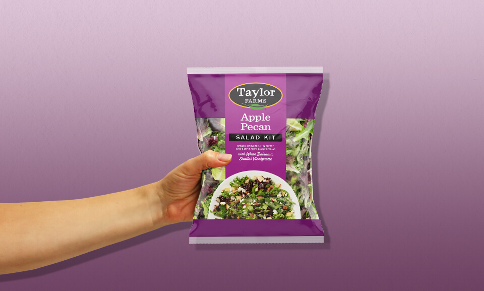 Hand holding a bag of Taylor Farms Apple Pecan Salad Kit showcasing fresh salad ingredients and packaging design.
