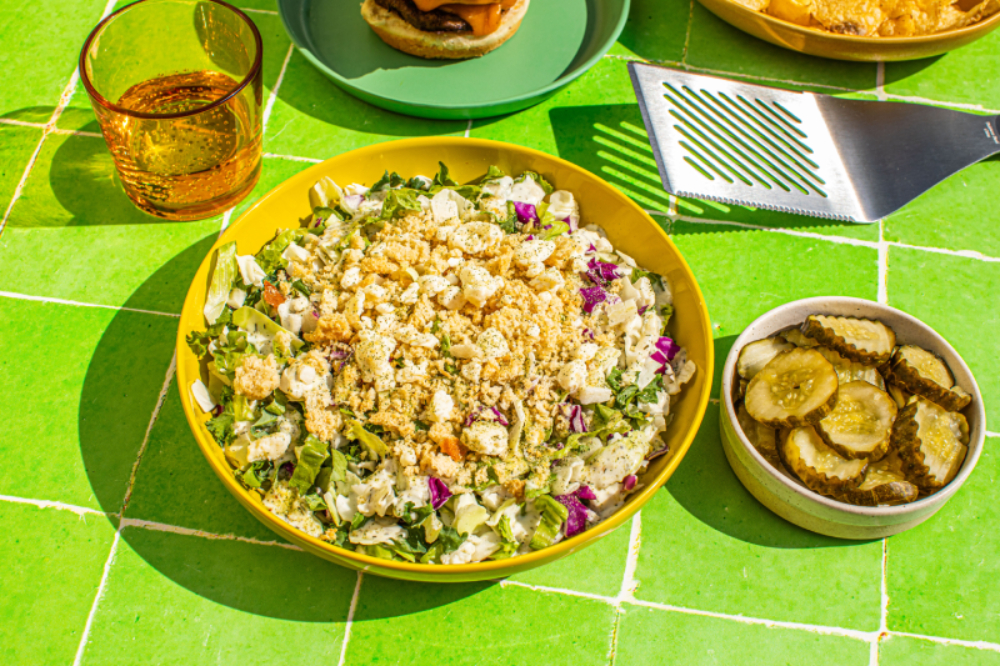 Taylor Farms Dill Pickle Chopped Salad in a Bowl around a Grill Scene