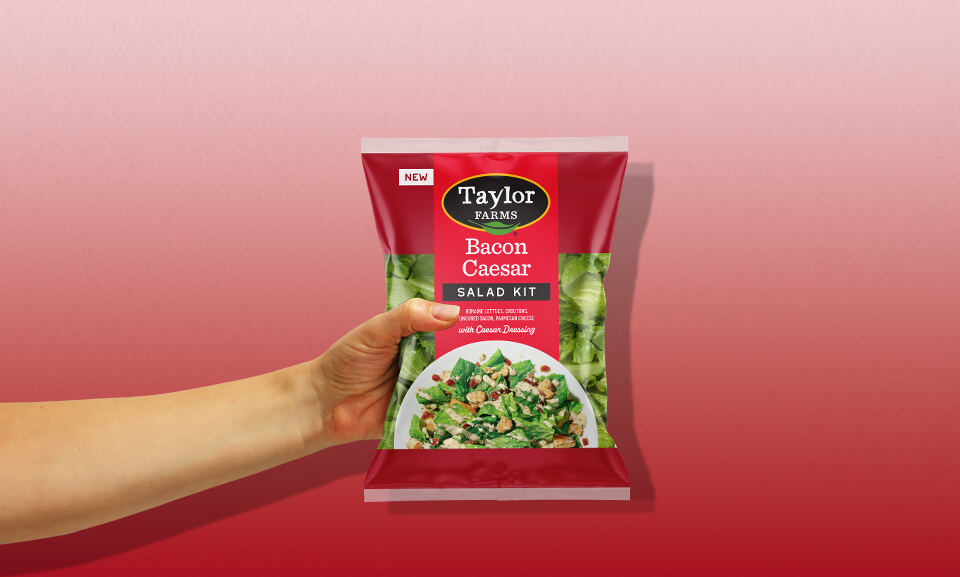 Hand holding a Bacon Caesar Salad Kit bag, showcasing fresh ingredients and convenient meal options from Taylor Farms.