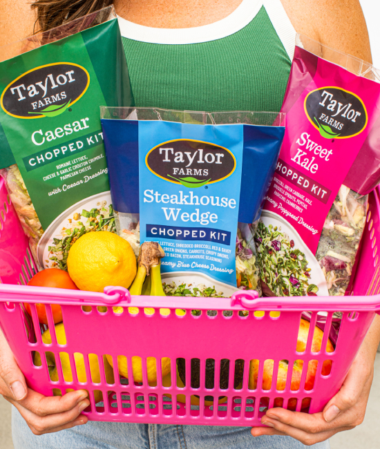 Three Taylor Farms chopped salad kits in a pink grocery basket with fruits and vegetables held by a woman in a green tank top