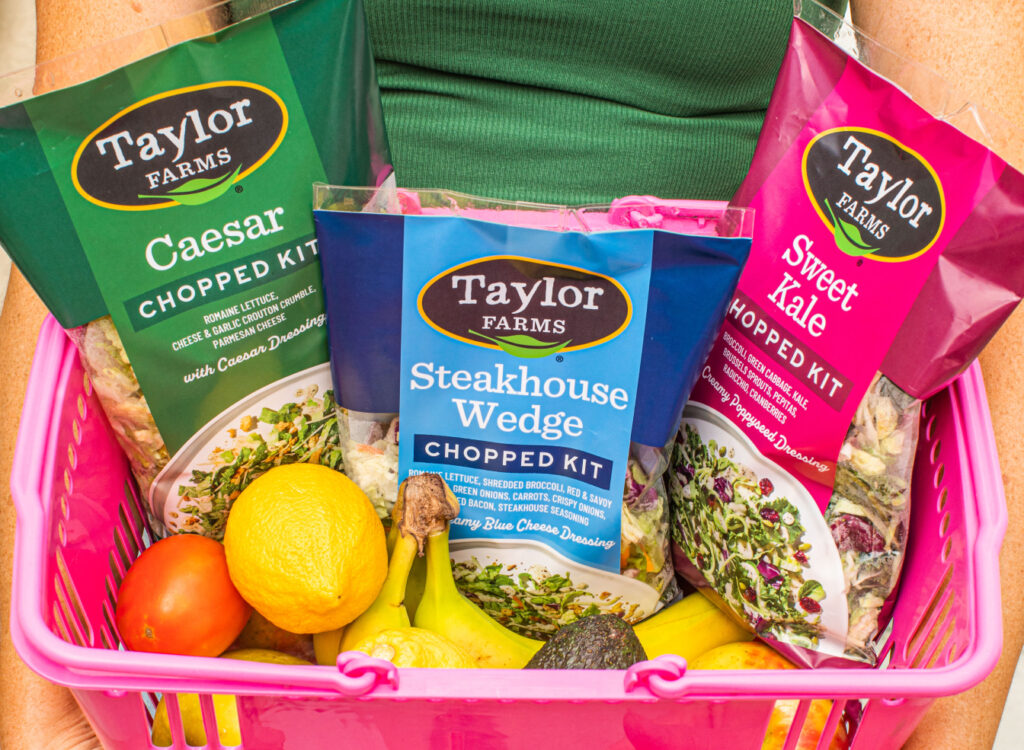 Three Taylor Farms chopped salad kits in a pink grocery basket with fruits and vegetables