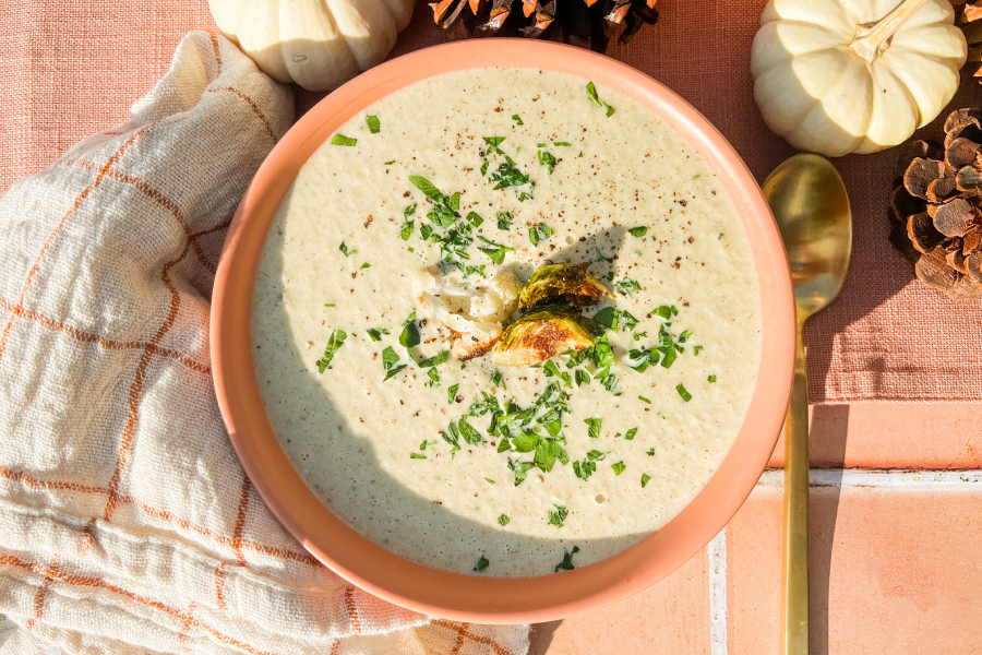 Roasted Cauliflower and Brussels Sprouts Soup in an orange bowl on a tiled table with a cloth napkin, small white pumpkins, and a gold spoon