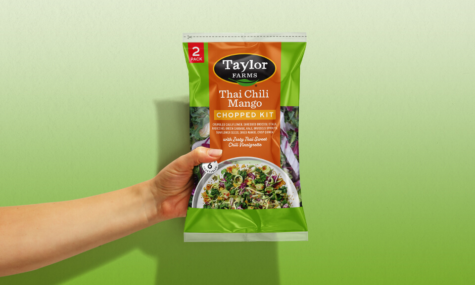 Hand holding a bag of Thai Chili Mango Chopped Salad Kit from Taylor Farms, showcasing vibrant packaging and fresh ingredients.